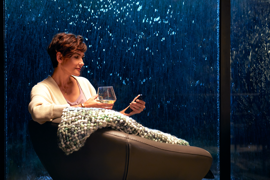 A woman relaxing in a chair with a glass of wine while using a smartphone to control smart home features, with illuminated water cascading down a glass wall behind her.